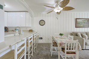 A kitchen with white cabinets, a dining table with chairs, a ceiling fan, and a clock on the wall. - Whispering Waters: A Tranquil One-bedroom Retreat With Peek Ocean Views (Oceanside)