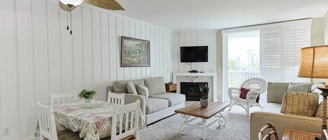 A living room with a white sofa, a dining table with chairs, a fireplace, and a television mounted on the wall.