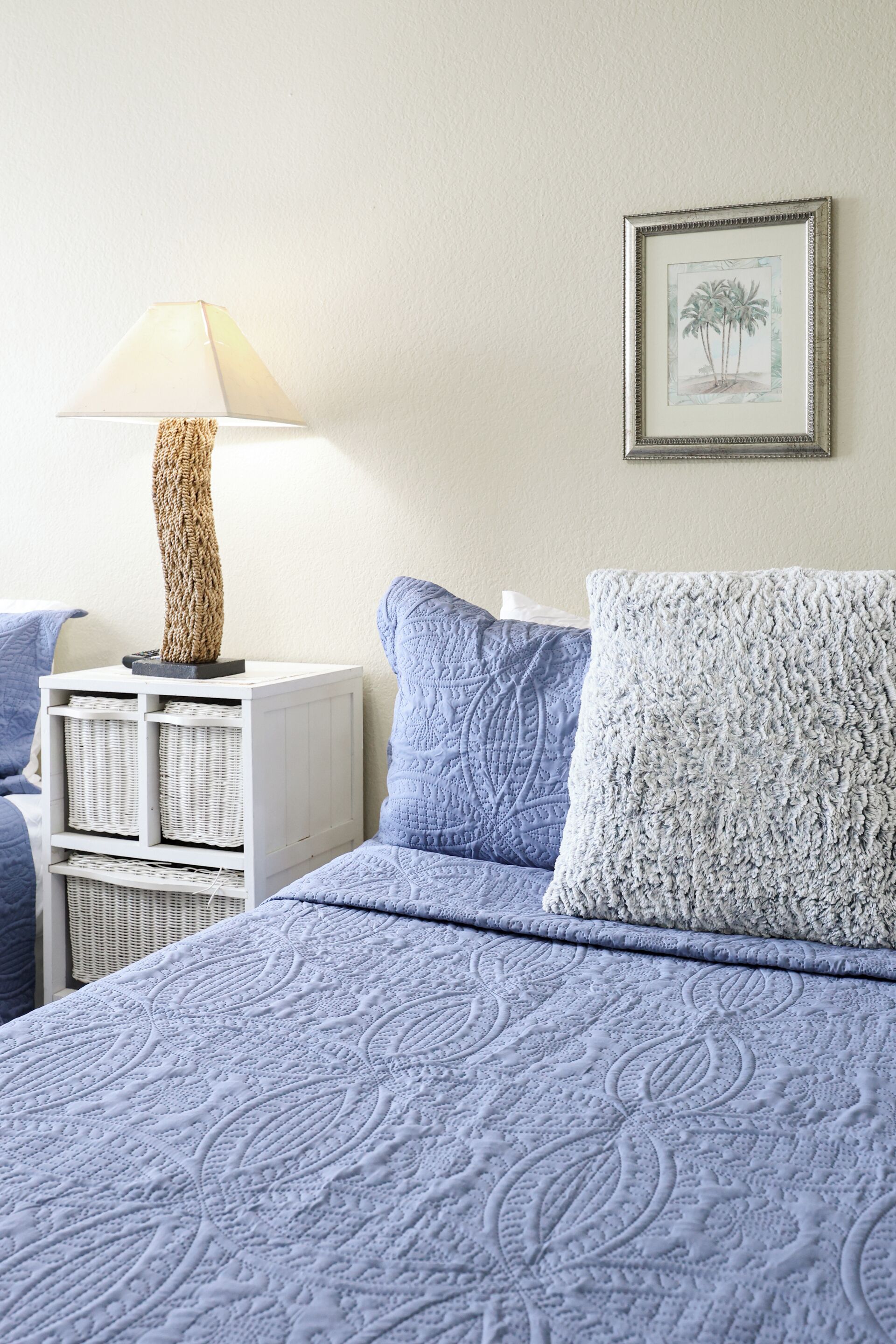 Un lit recouvert d’une couette bleue, une table de chevet blanche avec une lampe, et un tableau encadré accroché au mur.