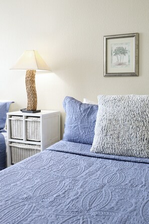 A bed with a blue quilt, a white nightstand with a lamp, and a framed picture on the wall. - Whispering Waters: A Tranquil One-bedroom Retreat With Peek Ocean Views (Oceanside)