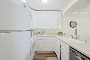 A kitchen with white cabinets, a dishwasher, a sink, and a clock on the wall. - Whispering Waters (Oceanside)