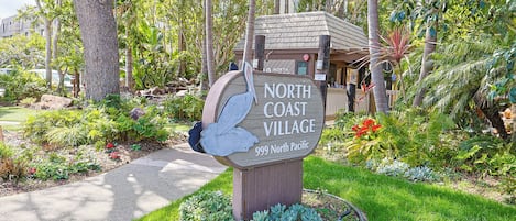A sign for North Coast Village, 999 North Pacific, surrounded by lush greenery and a well-maintained garden.