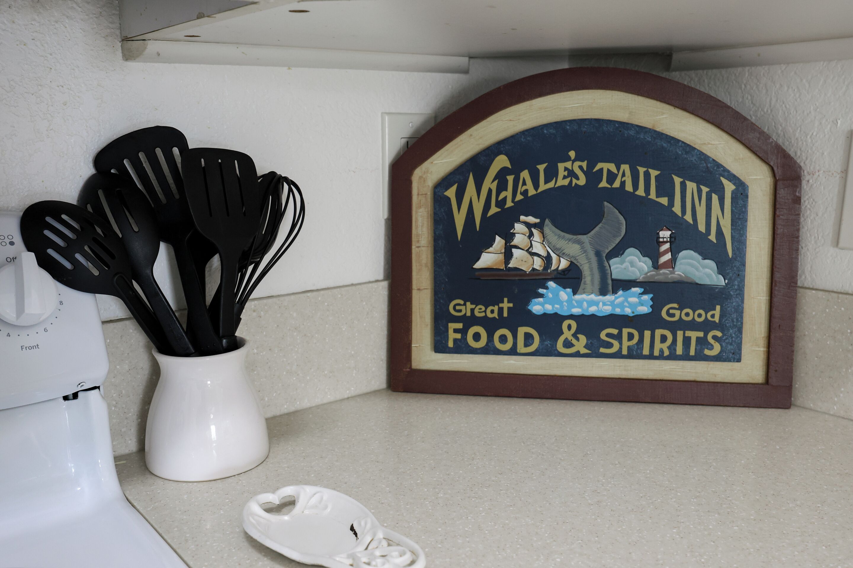 Un comptoir de cuisine sur lequel est posée une enseigne indiquant « Whale's Tail Inn », ainsi qu'un vase blanc contenant des spatules noires.