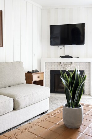 Ein Wohnzimmer mit einer Couch, einem Fernseher über einem Kamin und einem Topf mit einer Pflanze auf Fliesenboden.