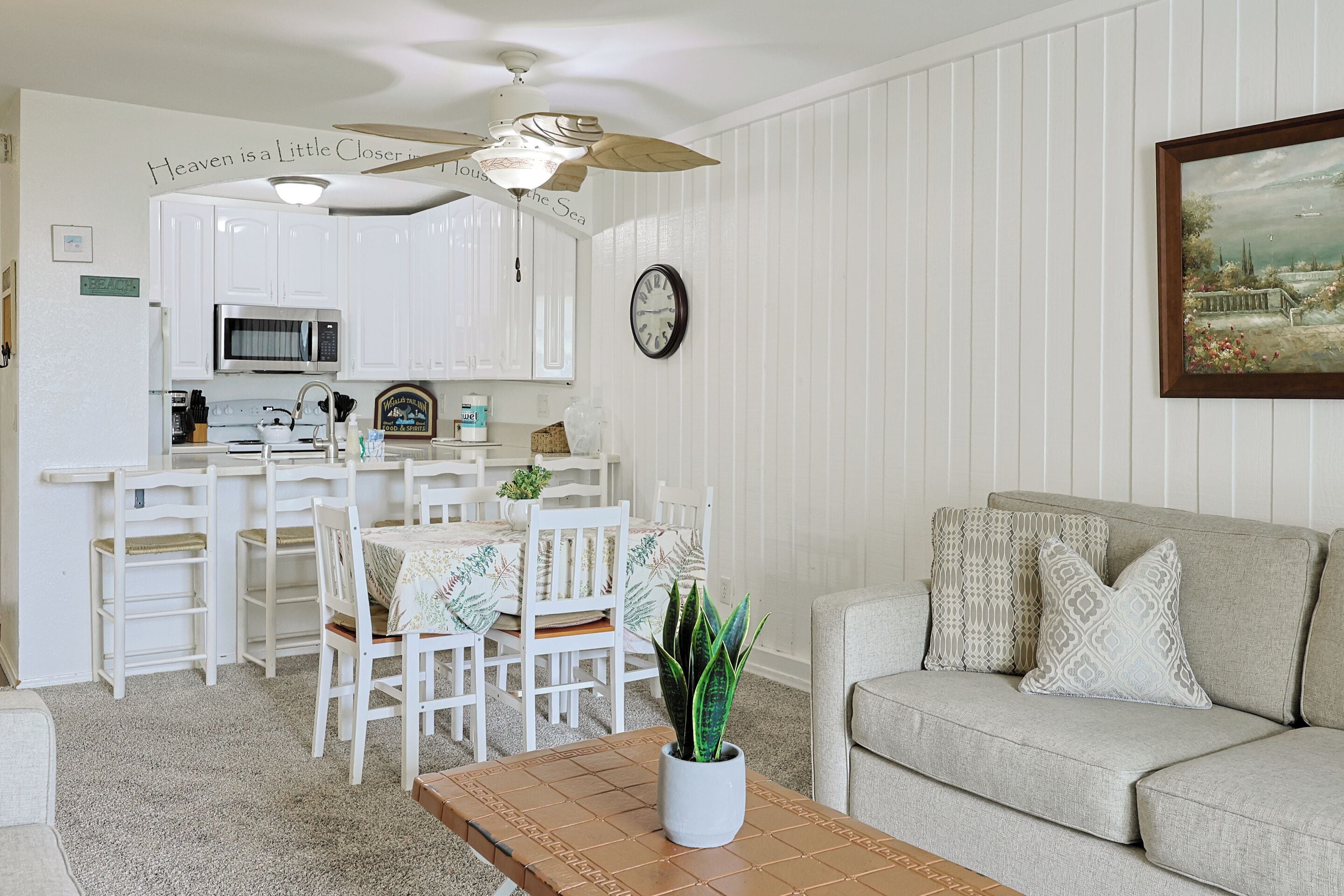 A kitchen with white cabinets, a microwave, and a ceiling fan. There is a dining table with chairs and a plant on it. A couch with patterned pillows is in the living area.