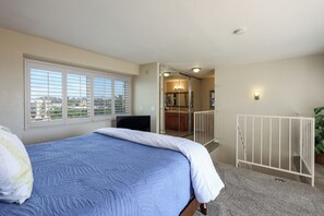 A bedroom with a large bed, a window with blinds, a white railing, and a view of the city. - Waves & Wonders (Oceanside)