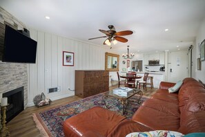 A living room with a brown leather sofa, a coffee table, and a patterned rug. There is a fireplace, a television, and a ceiling fan. The kitchen is visible in the background with white cabinets and a microwave. - Treetop Hideaway (Oceanside)