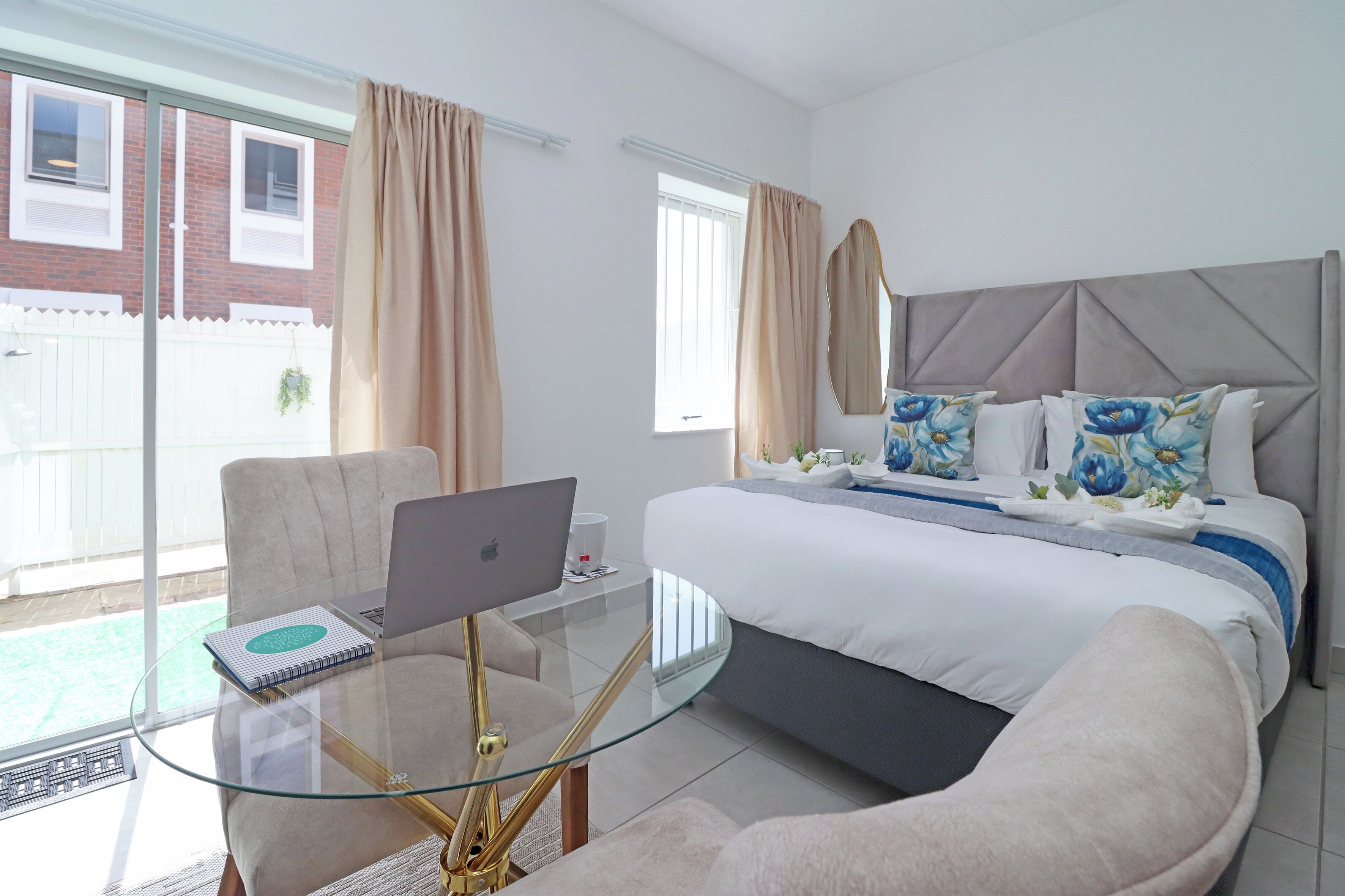 A modern bedroom with a glass table, a laptop, a bed with floral pillows, and a view of a brick building outside.