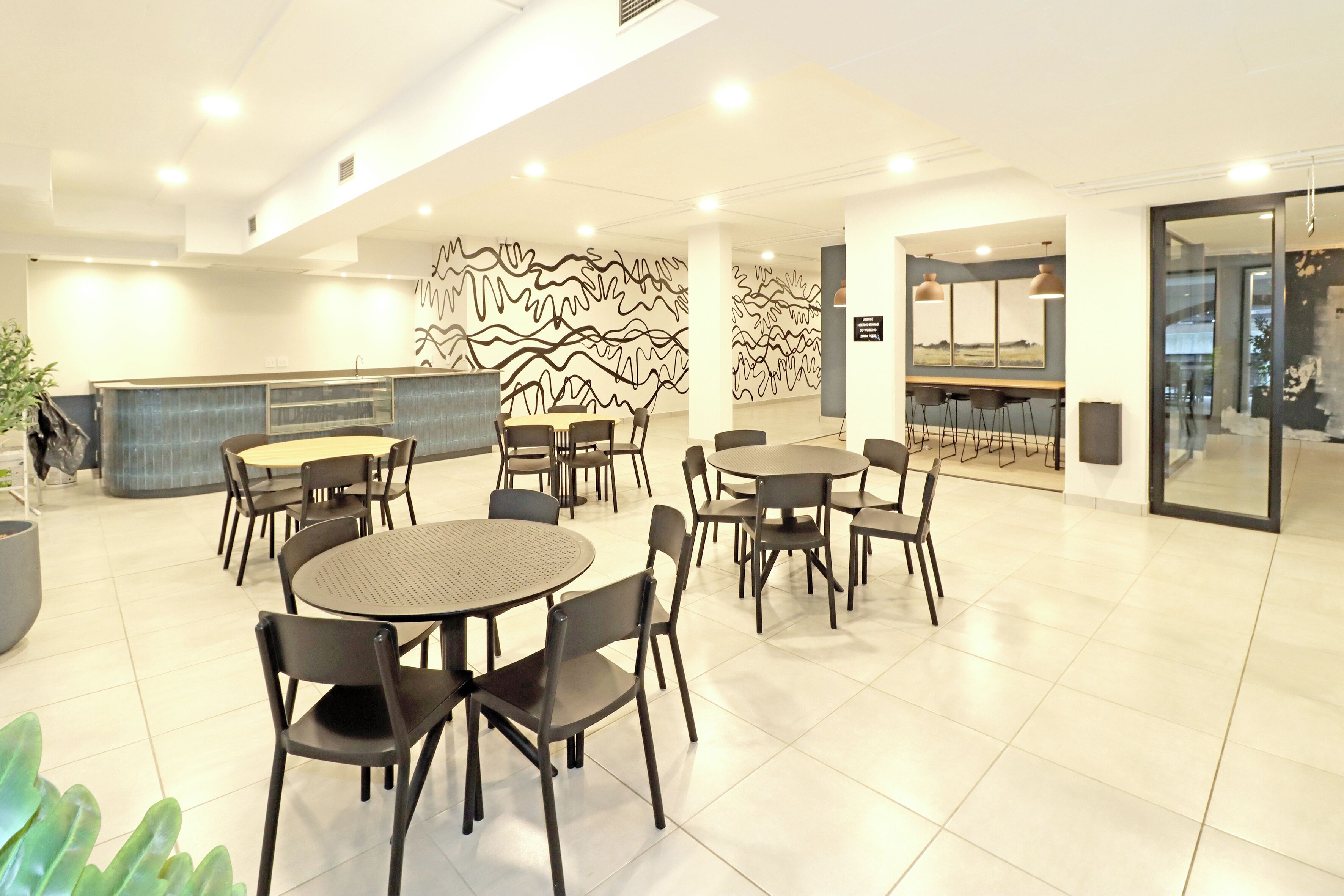A modern hotel lobby with round tables and chairs, a reception desk, and a decorative wall.