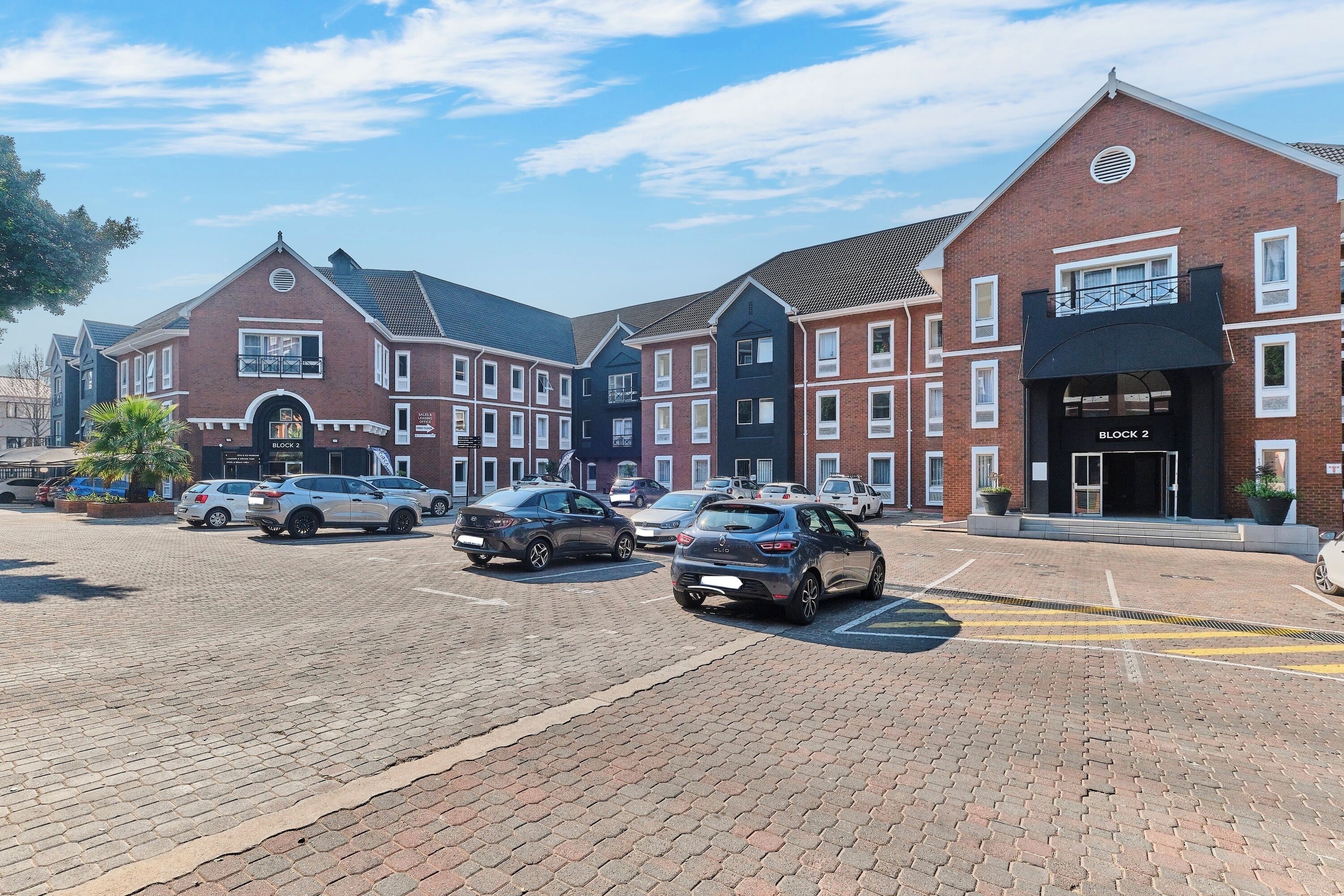 A parking area with multiple cars, a brick building with a sign for 'BLOCK 2', and a modern entrance with a sign 'BLOCK 2'.
