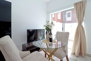 Een moderne woonkamer met een salontafel met glazen blad, twee fauteuils, een flatscreen televisie, een vaas met bloemen en een fles wijn.