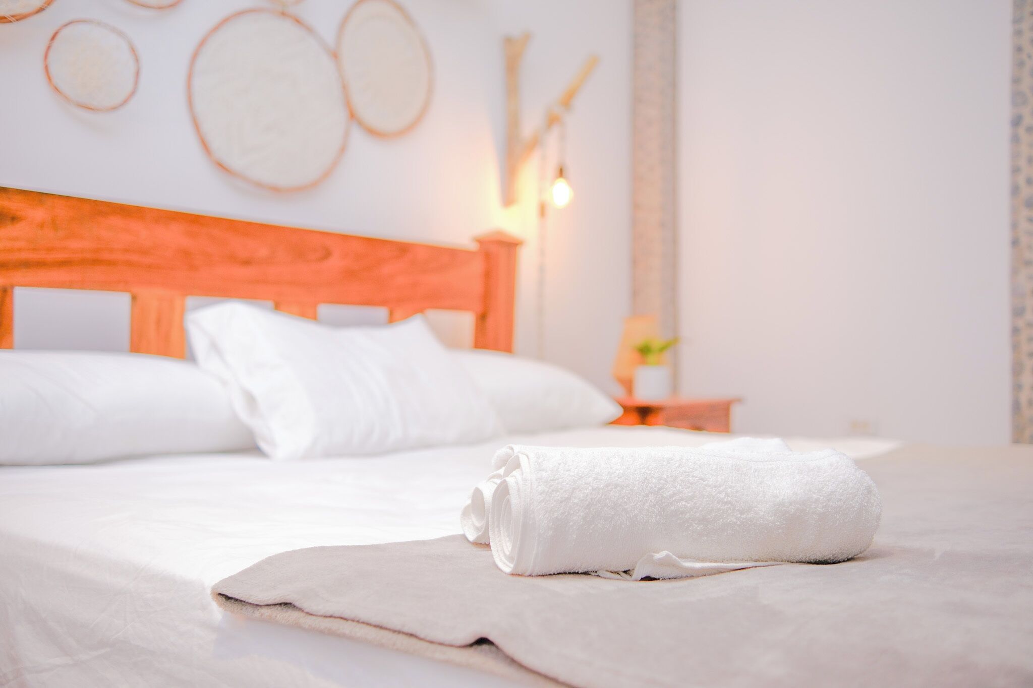 A neatly made bed with white linens and a rolled towel, a wooden headboard, and a bedside table with a lamp.