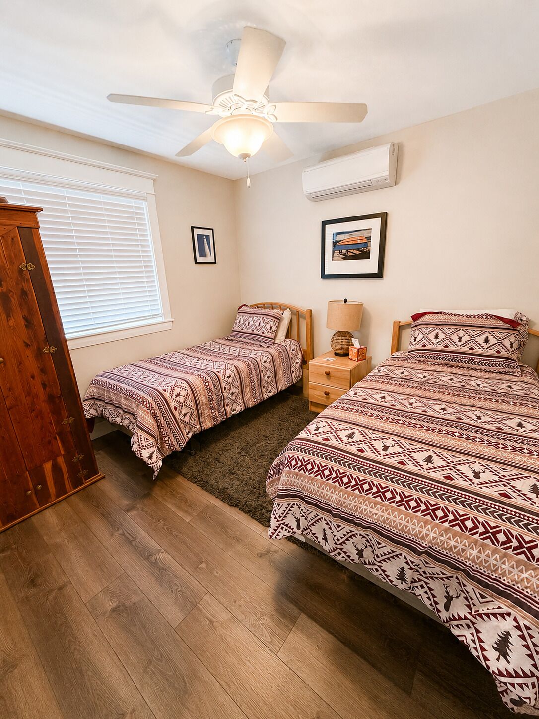 A bedroom with two beds, a ceiling fan, an air conditioning unit, a window with blinds, and a wooden wardrobe.