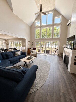 A spacious living room with a high ceiling, large windows, a fireplace, and a dining area with a view of the outdoors. - Sebago Lake Estate, 300’ Frontage, Inside the State Park, Private Dock & Kayaks (Casco)