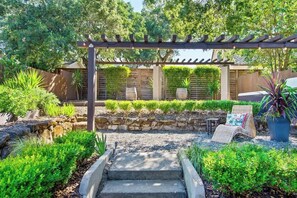 Ein Garten mit Terrassengarten, einer Pergola, einer Sitzgelegenheit und einem gepflasterten Weg zum Eingang.