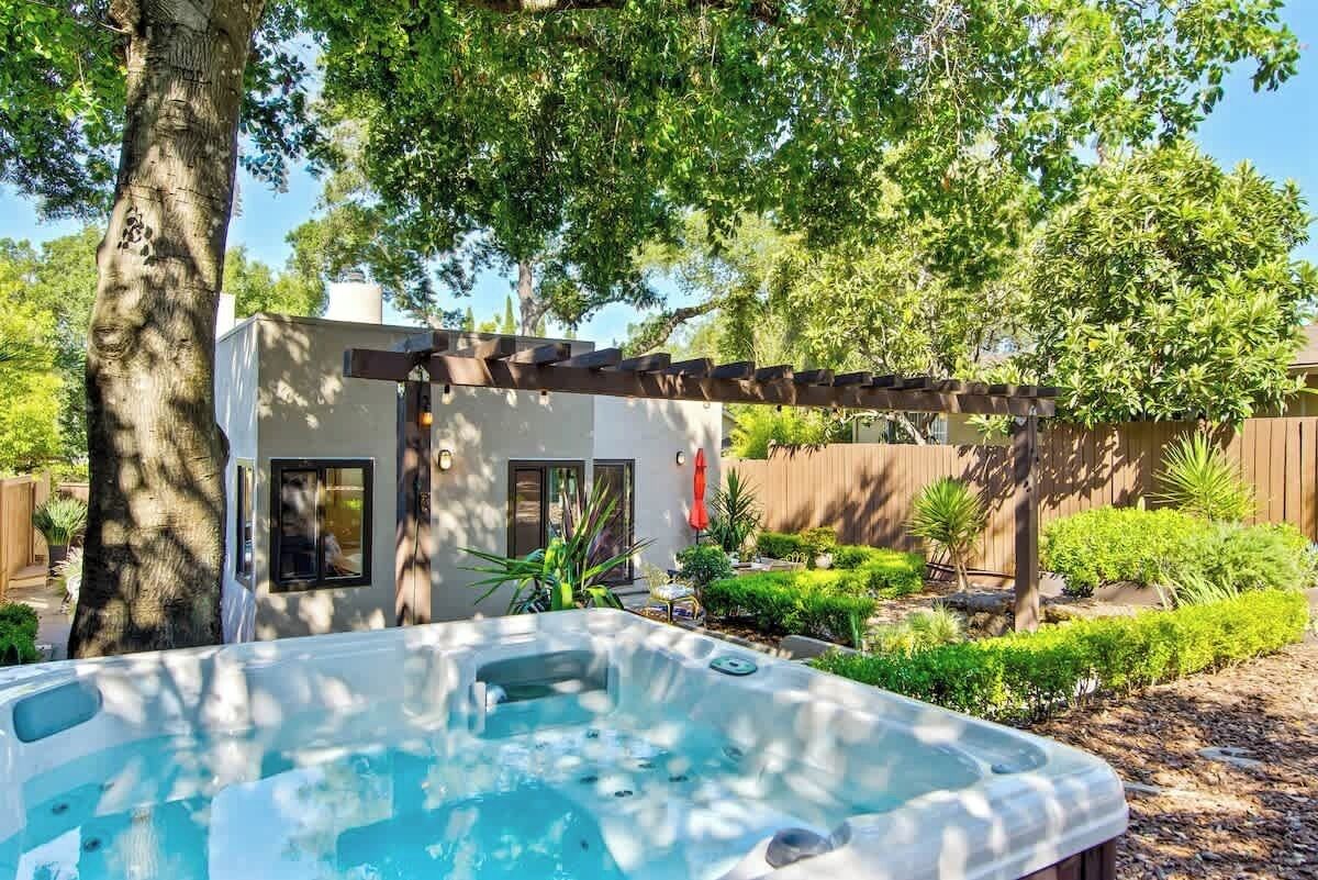 A hot tub in a backyard with a pergola, a small building, and various plants.