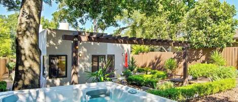 A hot tub in a backyard with a pergola, a small building, and various plants.