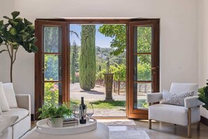A room with a large glass door leading to an outdoor garden, a white sofa, a white armchair, and a small table with a plant and wine glasses. - Hagen Heights Vineyard Estate Napa Valley (Napa)