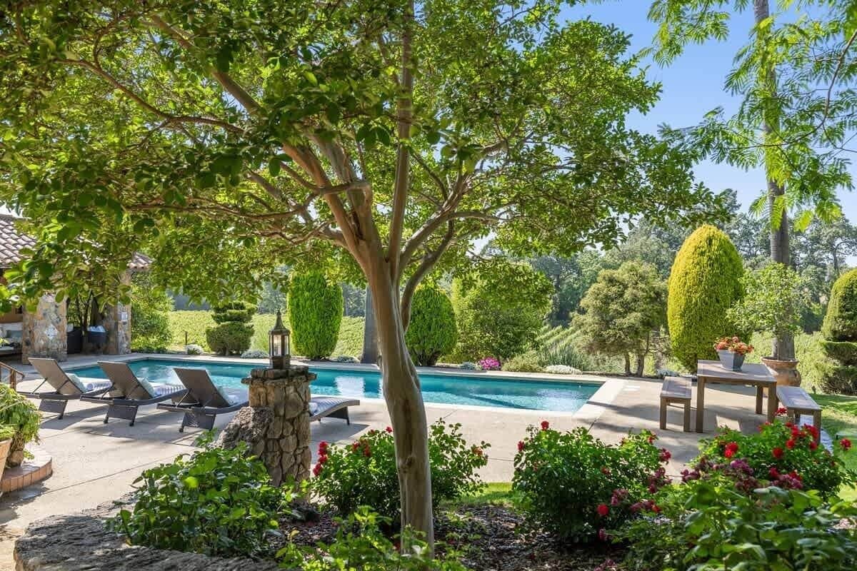 Un área junto a la piscina con sillones de descanso, un pilar de piedra y arbustos cuidados.