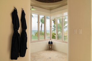 Ein Zimmer mit großem Fenster, das auf Palmen und Meer blickt, einer Duschbrause und einem kleinen Tisch mit Flaschen.