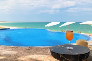 Ein Poolbereich mit freiem Blick auf das Meer, ein Getränk auf einem Tisch und ein Strohhut.