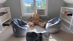 Una habitación con un ventanal grande, dos sillones tipo puffs, un osito de peluche y estanterías con libros y juegos.