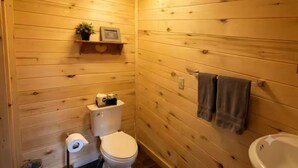 A wooden-paneled bathroom with a toilet, a shelf with a plant and a framed item, and two towels hanging on the wall. - Cabin#5 in Lake George Cabins & RV Park (Lake George)