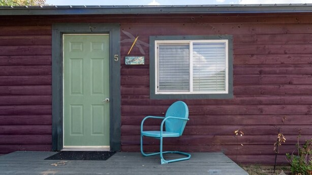 A wooden cabin with a green door, a window with blinds, and a blue chair on the porch. - Cabin#5 in Lake George Cabins & RV Park (Lake George)