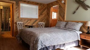 A bedroom with a bed, two chairs, a nightstand, and a wooden wall decoration. - Cabin#5 in Lake George Cabins & RV Park (Lake George)