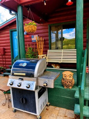 A barbecue grill on a porch with a bench and an owl decoration. - Cabin#6 in Lake George Cabins & RV Park (Lake George)