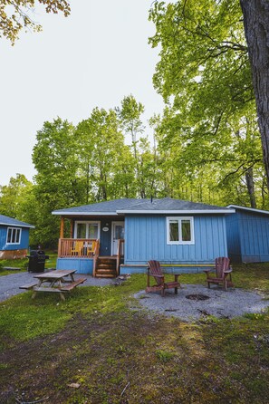 A small cabin with a porch, a picnic table, and two Adirondack chairs. - Pelican Cottage - 2-Bedroom Cottage – Family Friendly (Trent Lakes)