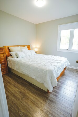 A bedroom with a wooden bed, a nightstand, a bedside lamp, and a window. - Pelican Cottage - 2-Bedroom Cottage – Family Friendly (Trent Lakes)