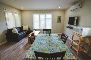 Ein Wohnzimmer mit einer schwarzen Ledersofa, einem Esstisch mit Stühlen, einem gemusterten Teppich, einem Fernseher und einer Mikrowelle.