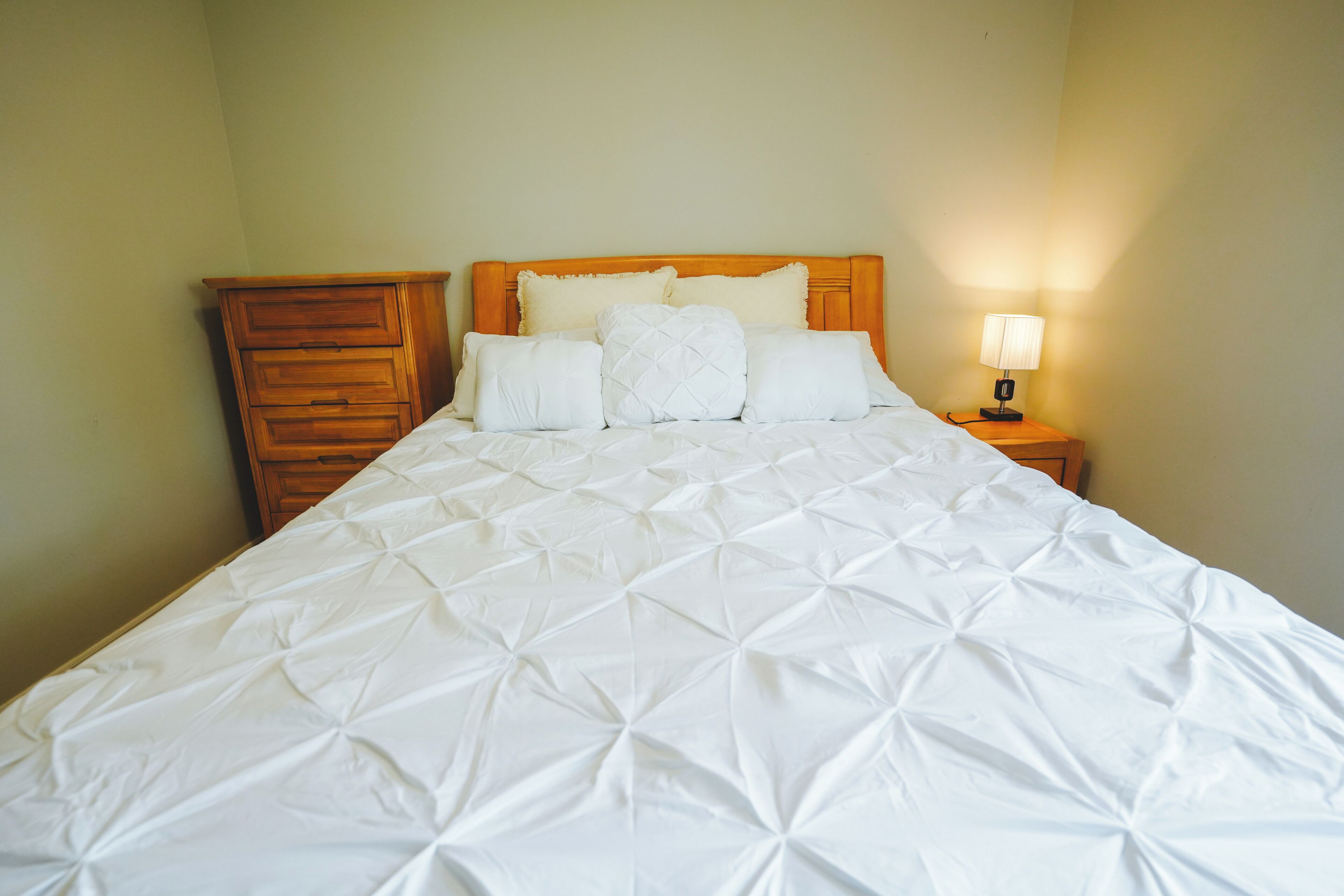 A neatly made bed with a white quilt, a wooden headboard, and a bedside lamp.