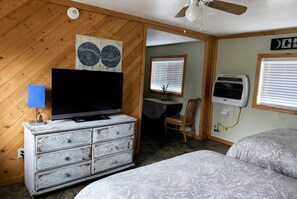 Quarto com televisão em uma cômoda, ventilador de teto, cama com roupa de cama estampada e escrivaninha com cadeira.