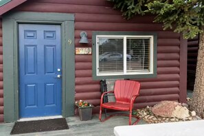A log cabin with a blue door, a window with blinds, a red chair, and a small porch area. - Cabin#9 in Lake George Cabins & RV Park (Lake George)