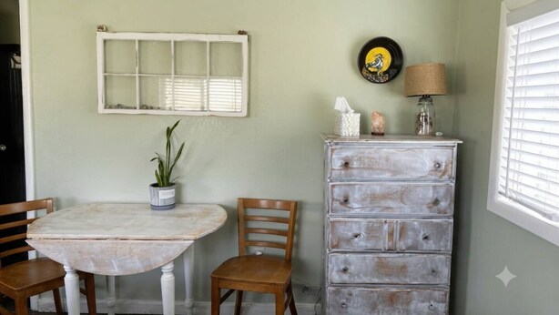 Un comedor con una mesa redonda, dos sillas, una planta en maceta, un aparador con cajones, un espejo y un reloj de pared.