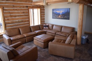 Interior de cabana de tronco com sofá seccional, apoio para os pés e um quadro de uma paisagem montanhosa.