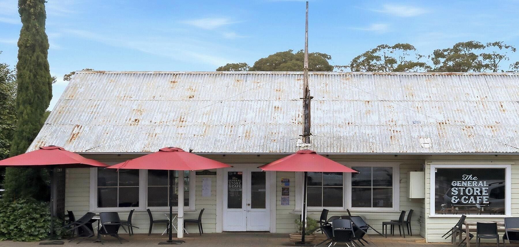 A building with a corrugated metal roof, outdoor seating with red umbrellas, and a sign for 'The General Store & Cafe.'