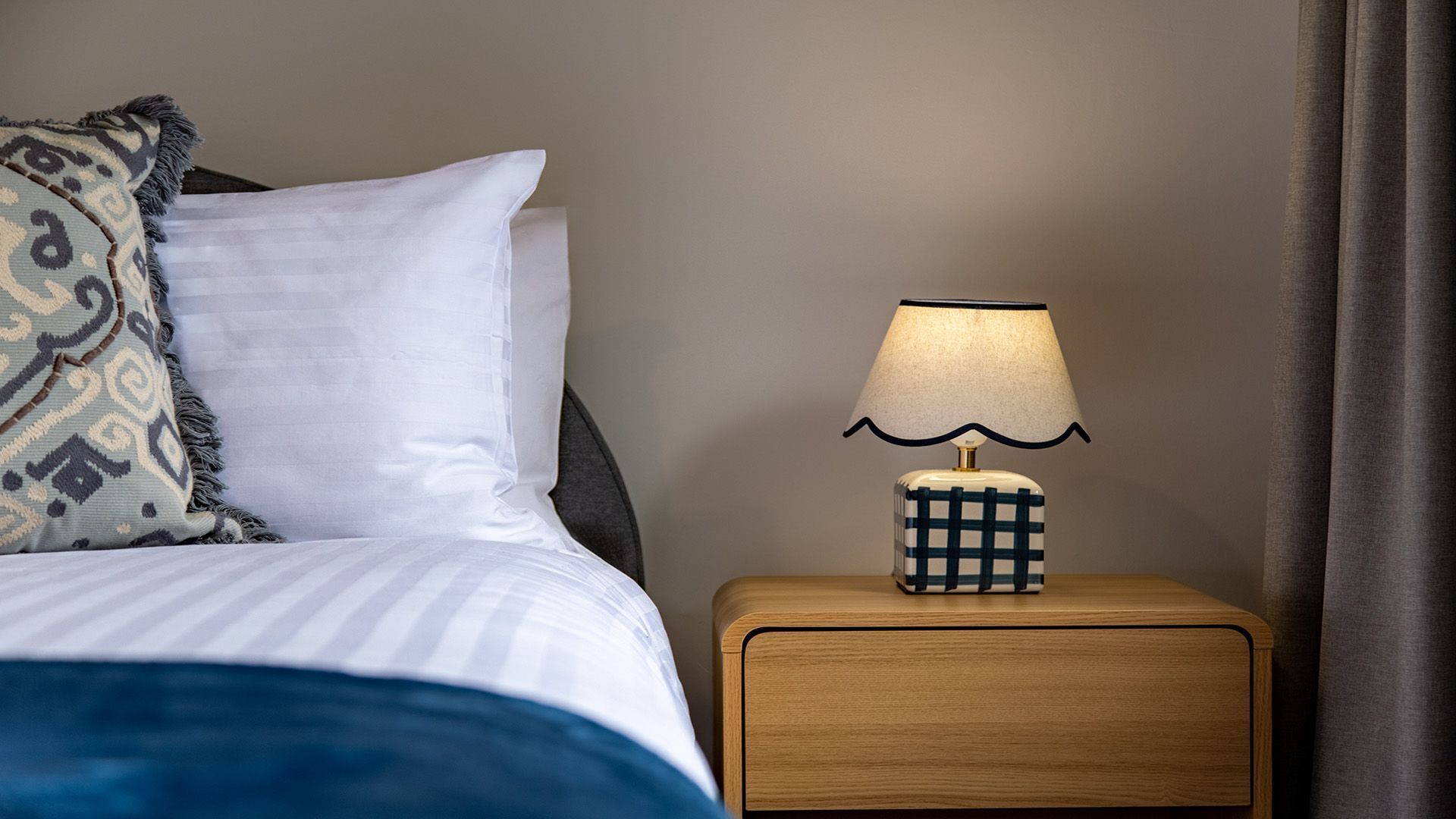 A neatly made bed with a pillow, a bedside table with a lamp, and a curtain on the right.