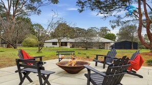An outdoor seating area with a fire pit, surrounded by trees and a house in the background.