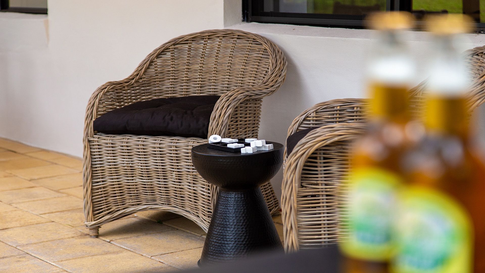 Wicker chairs with cushions, a small table with a black base, and a bottle in the foreground.