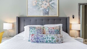 Una cama bien hecha con un cabecero tapizado gris, dos almohadas blancas y un colorido edredón bordado. Encima de la cama hay una obra de arte floral enmarcada.