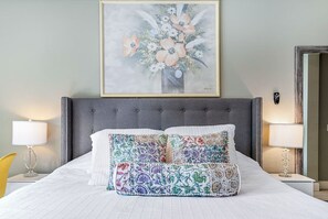 En snyggt bäddad säng med en grå klädd sänggavel, två vita kuddar och en färgglad, broderad pläd. Ovanför sängen hänger ett inramat konstverk med blommotiv.