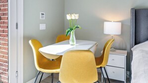 Habitación de hotel con una mesa blanca, sillas amarillas, un jarrón con flores blancas, una lámpara de noche y una cama con cabecera gris.
