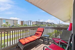 A balcony with two lounge chairs, one with a red cushion and another with a grey cushion, overlooking a street with buildings in the background. - Coastal Getaway on Okaloosa Island - 3 Minute Walk to Beach! (Fort Walton Beach)
