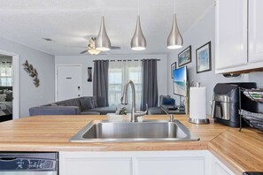 A modern kitchen with a central sink, overhead pendant lights, and a dining area with a sofa and a coffee machine. - Coastal Getaway on Okaloosa Island - 3 Minute Walk to Beach! (Fort Walton Beach)