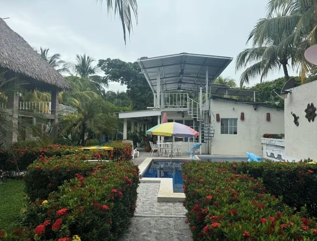 A pool area with a thatched-roof structure, a covered seating area, and a white building with a satellite dish.