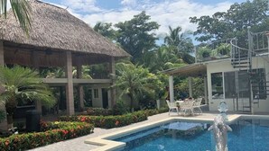 A swimming pool with a dolphin statue, surrounded by tropical plants and a thatched-roof structure.
