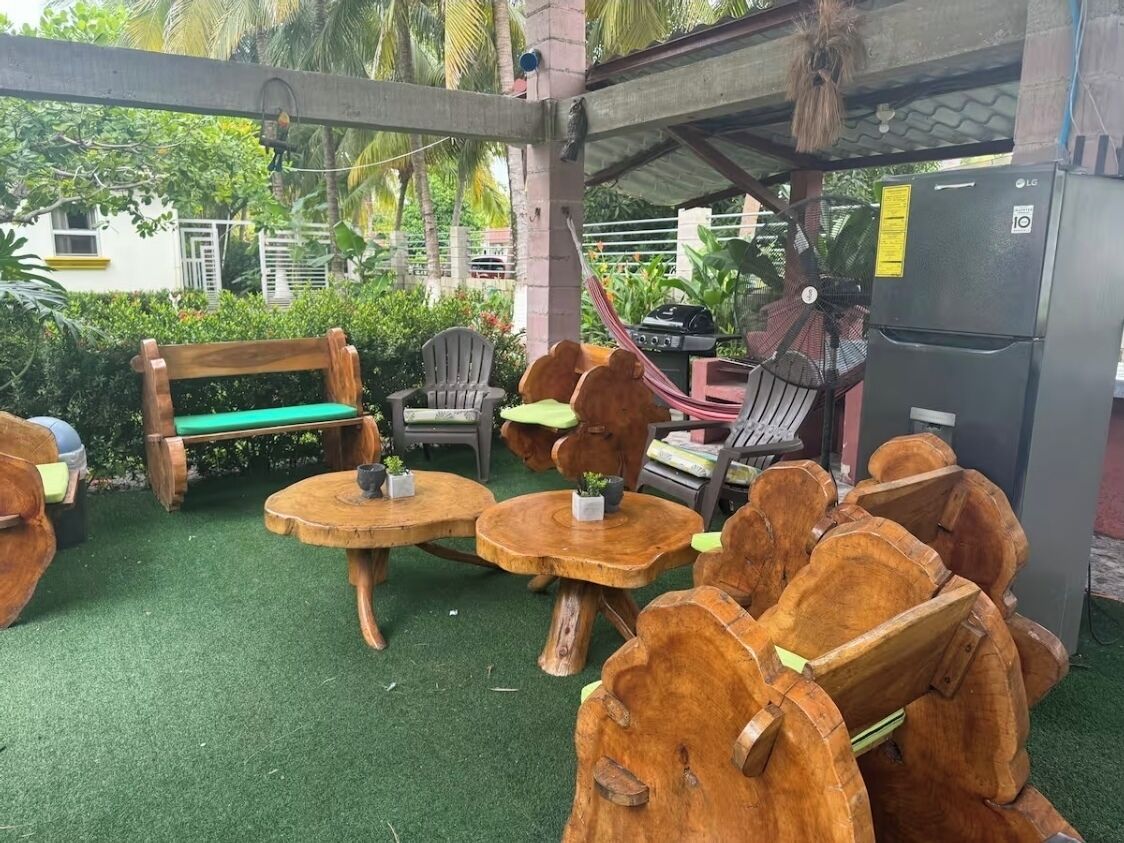 A covered outdoor seating area with wooden furniture, a grill, and a refrigerator.